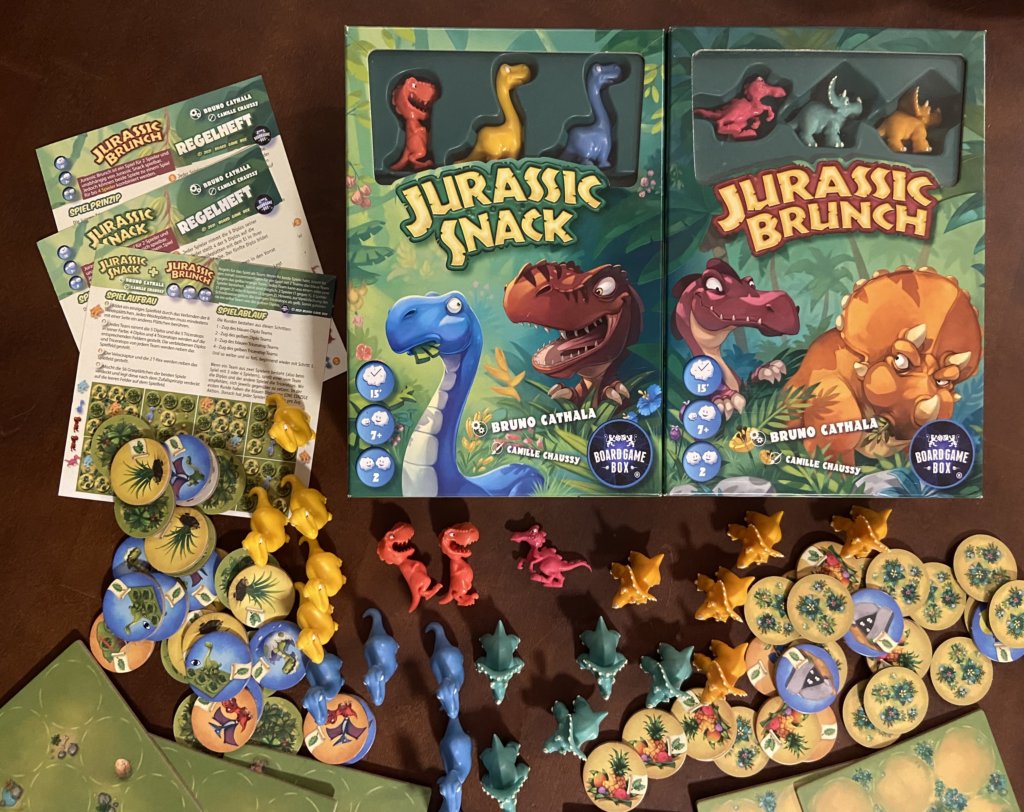 Figuren und Plättchen und die Schachteln der Spiele Jurassic Snack und Jurassic Brunch