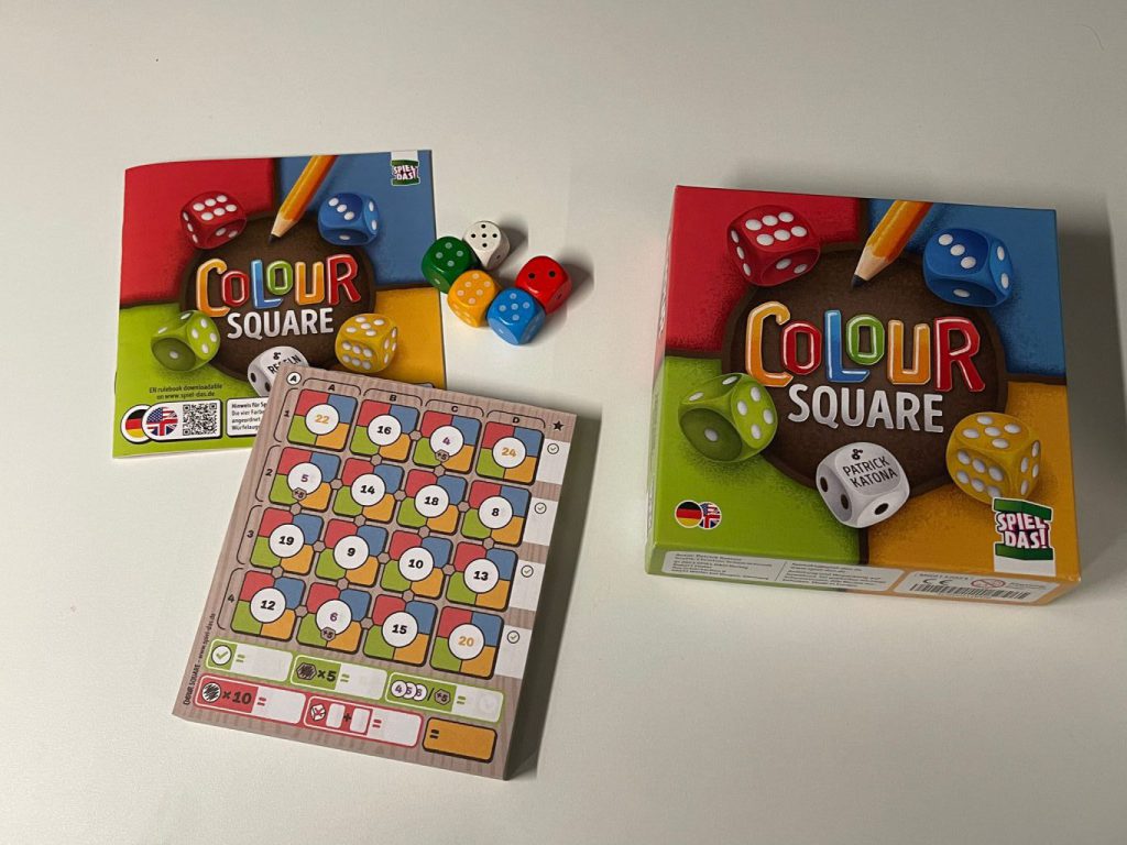 Das Bild enthält die Box vom Spiel Colour Square und die Materialien, mit den Würfeln, dem Spielblock und der Anleitung.