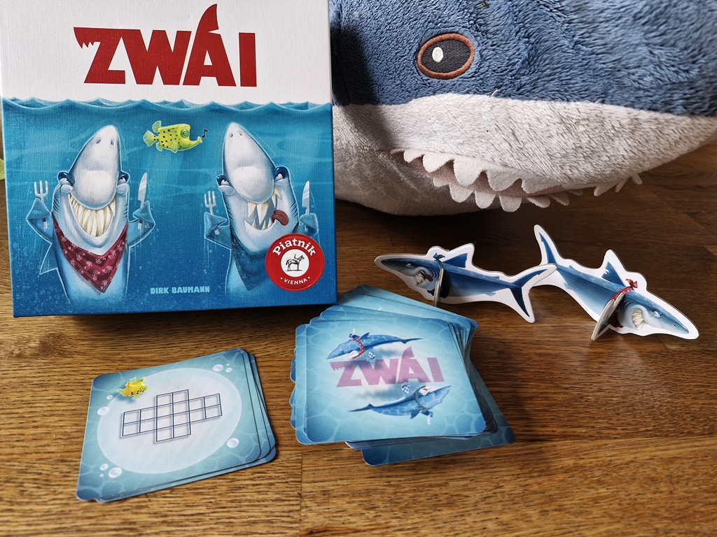 Spielkarton und Inhalt, im Hintergrund ein Plüsch-Hai